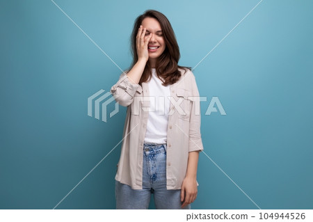 modest brunette young female adult in a shirt and jeans covered her face with her hand on a blue modest brunette young female adult in a shirt and jeans covered her face with her hand on a blue 104944526