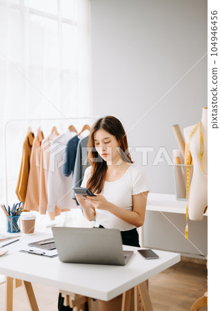 Asian young woman on desk in office of fashion designer and holds tablet, laptop and smartphone on white table 104946456