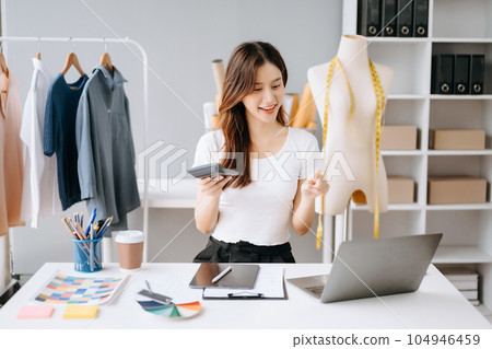Asian young woman on desk in office of fashion designer and holds tablet, laptop and smartphone on white table 104946459