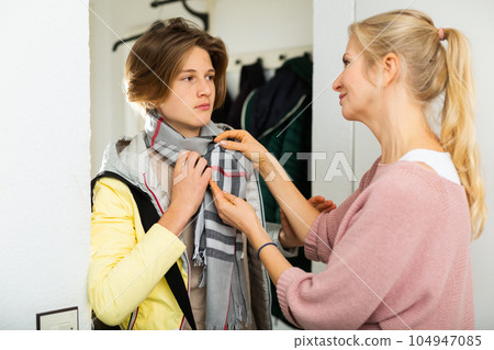 Caring woman seeing out teenage son ready to go to school, straightening his scarf in doorway at home 104947085