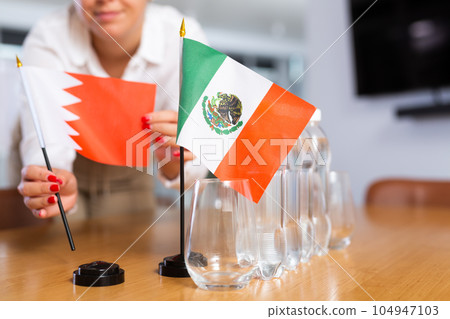 A woman in a white blouse with friendly smile arrangs flags of Mexico and Bahrain for meeting. 104947103