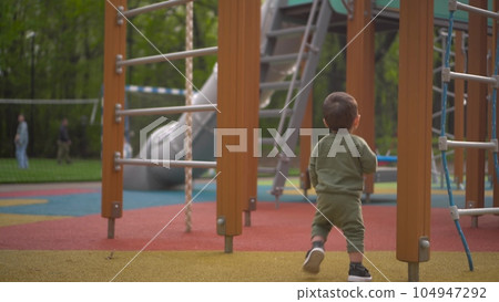 A two-year-old child walks on a playground among slides and swings. Little son in the park. 104947292