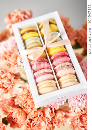 Dessert French Macarons in present box on a background of flowers. Sweet dessert on a flowers background. Still life with macaroni cakes and different flower. Delivery concept, coffee and flower shop 104947361