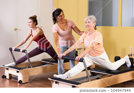 Elderly woman practicing pilates on reformer with Hispanic female trainer 104947944