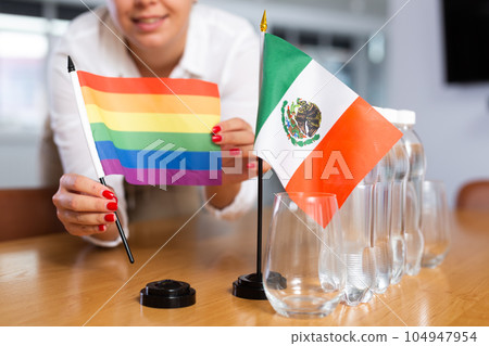 A woman in a white blouse with friendly smile arrangs flags of Mexico and LGBT for meeting. A woman in a white blouse with friendly smile arrangs flags of Mexico and LGBT for meeting. 104947954