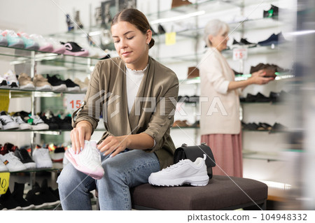 In shoe store,woman sits on ottoman and tries on sneakers 104948332
