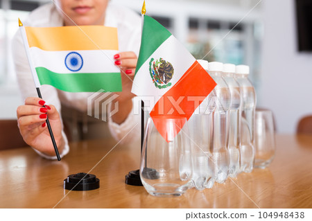 A woman in a white blouse with friendly smile arrangs flags of Mexico and Iran for meeting. 104948438