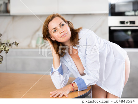 Woman in unbuttoned white shirt is standing in kitchen and leaning elbows on dining table 104948584