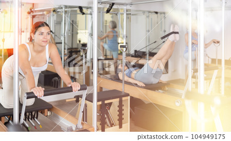 Positive young girl practicing pilates stretching exercises on reformer at gym Positive young girl practicing pilates stretching exercises on reformer at gym 104949175