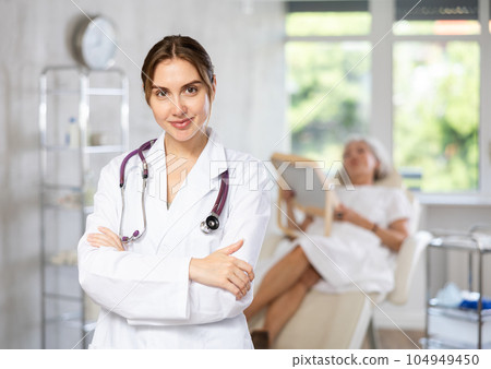 Smiling senior woman doctor in uniform with woman patient lying behind in clinic. Smiling senior woman doctor in uniform with woman patient lying behind in clinic. 104949450