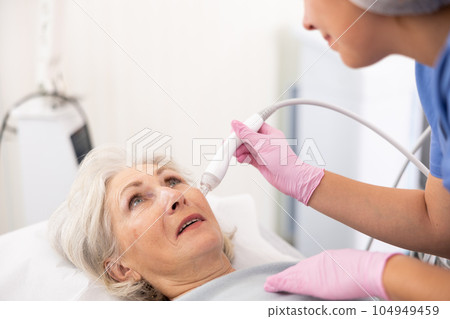 Elderly woman at appointment with beautician doing ultrasonic face cleaning with help of high-frequency ultrasonic waves in cosmetology clinic Elderly woman at appointment with beautician doing ultrasonic face cleaning with help of high-frequency ultrasonic waves in cosmetology clinic 104949459