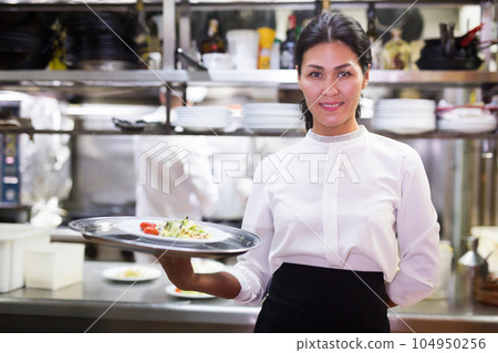 portrait of smiling female waiter standing in kitchen in restaurante 104950256