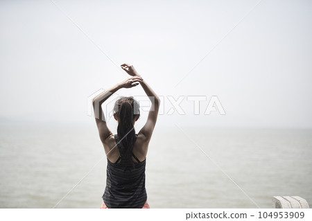 young asian adult woman stretching arms outdoors by the sea, rear view 104953909