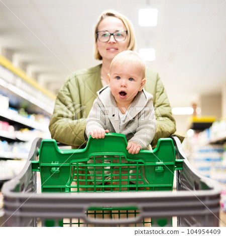 Mother pushing shopping cart with her infant baby boy child down department aisle in supermarket grocery store. Shopping with kids concept Mother pushing shopping cart with her infant baby boy child down department aisle in supermarket grocery store. Shopping with kids concept 104954409