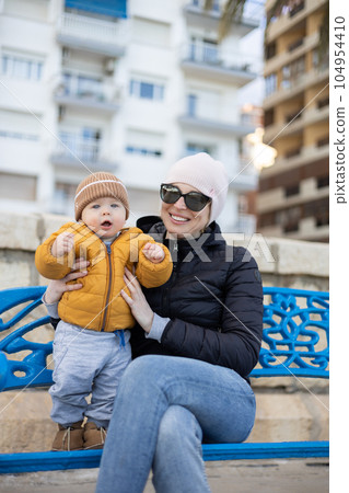 Young mother with her cute infant baby boy child on bench in city park Young mother with her cute infant baby boy child on bench in city park 104954410