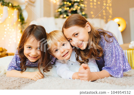 two girls and a little boy lie on the floor in a room with Christmas decor. 104954532