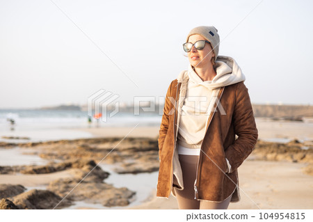 Portrait of young stylish woman wearing brown padded jacket, hoodie, wool cap and sunglasses on long sandy beach in spring 104954815