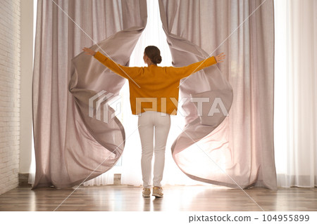 Woman opening window curtains at home in morning, back view Woman opening window curtains at home in morning, back view 104955899