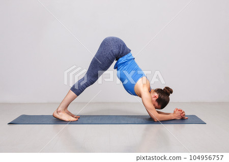 Woman doing Hatha yoga Surya Namaskar Sun Salutation asana Adhomukha svanasana variation - Ardha Pincha Mayurasana dolphin pose on yoga mat in studio on grey bagckground 104956757