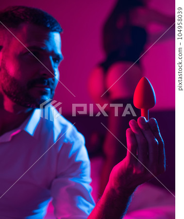 Bearded caucasian man holding an anal plug. Close-up of female buttocks in underwear. Sexual preferences of the couple. 104958039