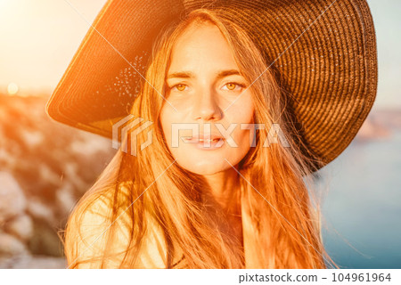 Portrait of happy young woman wearing summer black hat with large brim at beach on sunset. Closeup face of attractive girl with black straw hat. Happy young woman smiling and looking at camera at sea. 104961964
