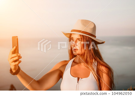 Woman travel sea. Happy tourist in hat enjoy taking picture outdoors for memories. Woman traveler posing on the beach at sea surrounded by volcanic mountains, sharing travel adventure journey. 104962050