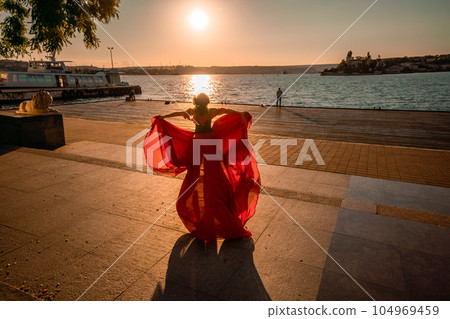 Sunrise red dress. A woman in a long red dress against the backdrop of sunrise, bright golden light of the sun's rays. The concept of femininity, harmony. 104969459