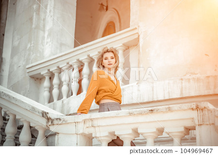 A middle-aged woman looks like a good blonde with curly beautiful hair and makeup on the background of the building. She is wearing a yellow sweater. 104969467