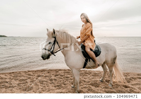 A white horse and a woman in a dress stand on a beach, with the sky and sea creating a picturesque backdrop for the scene. A white horse and a woman in a dress stand on a beach, with the sky and sea creating a picturesque backdrop for the scene. 104969471