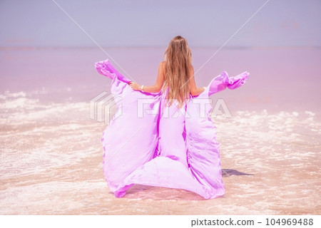 Woman pink salt lake. Against the backdrop of a pink salt lake, a woman in a long pink dress takes a leisurely stroll along the white, salty shore, capturing a wanderlust moment. 104969488