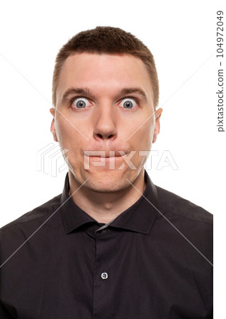Handsome young man in a black shirt is making faces, while standing isolated on a white background Handsome young man in a black shirt is making faces, while standing isolated on a white background 104972049