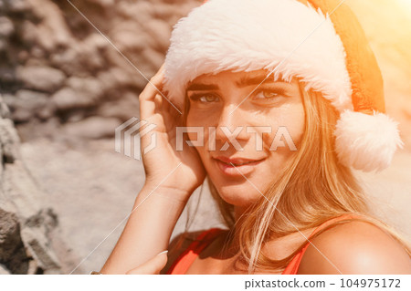 Woman summer travel sea. Happy tourist in red bikini and Santas hat enjoy taking picture outdoors for memories. Woman traveler posing on the beach surrounded by volcanic mountains, sharing travel joy Woman summer travel sea. Happy tourist in red bikini and Santas hat enjoy taking picture outdoors for memories. Woman traveler posing on the beach surrounded by volcanic mountains, sharing travel joy 104975172