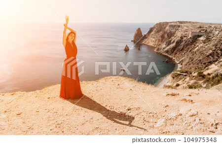 Woman in red dress on sea. Side view a Young beautiful sensual woman in a red long dress posing on a rock high above the sea on sunset. Girl on the nature on blue sky background. Fashion photo. 104975348
