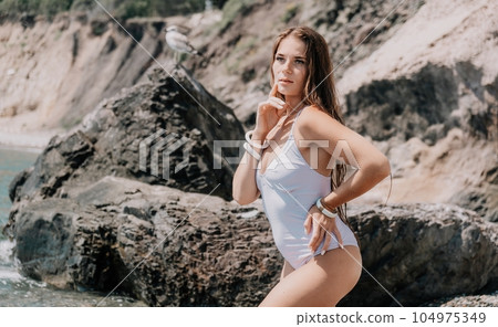 Woman travel sea. Happy tourist in white bikini enjoy taking picture outdoors for memories. Woman traveler posing on the beach at sea surrounded by volcanic mountains, sharing travel adventure journey 104975349