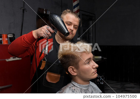 A man, a professional hairdresser, Caucasian, stylish dries the hair of a client, a guy with a hairdryer in the process of cutting at work in the salon A man, a professional hairdresser, Caucasian, stylish dries the hair of a client, a guy with a hairdryer in the process of cutting at work in the salon 104975500