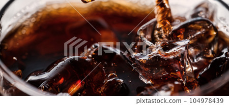 Pouring of Cola and Ice. Cola soda and ice splashing fizzing or floating up to top of surface. Close up of ice in cola water. Texture of carbonate drink with bubbles in glass. Cold drink background Pouring of Cola and Ice. Cola soda and ice splashing fizzing or floating up to top of surface. Close up of ice in cola water. Texture of carbonate drink with bubbles in glass. Cold drink background 104978439