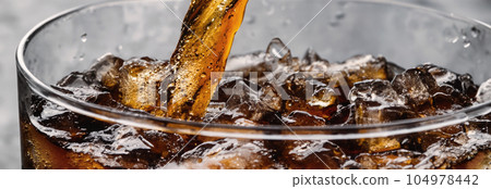 Pouring of Cola and Ice. Cola soda and ice splashing fizzing or floating up to top of surface. Close up of ice in cola water. Texture of carbonate drink with bubbles in glass. Cold drink background Pouring of Cola and Ice. Cola soda and ice splashing fizzing or floating up to top of surface. Close up of ice in cola water. Texture of carbonate drink with bubbles in glass. Cold drink background 104978442