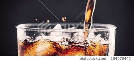 Pouring of Cola and Ice. Cola soda and ice splashing fizzing or floating up to top of surface. Close up of ice in cola water. Texture of carbonate drink with bubbles in glass. Cold drink background Pouring of Cola and Ice. Cola soda and ice splashing fizzing or floating up to top of surface. Close up of ice in cola water. Texture of carbonate drink with bubbles in glass. Cold drink background 104978445