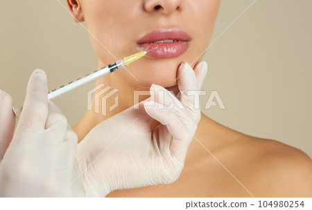Woman getting lip injection on beige background, closeup Woman getting lip injection on beige background, closeup 104980254