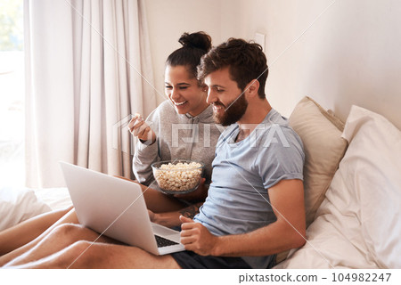 Watching movie, popcorn and couple in bed with laptop for streaming, food and internet. Subscription, happy and comic with man and woman at home for social media, funny and free time on weekend 104982247