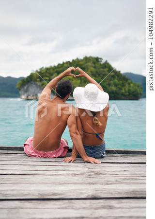 Heart shape, hands and couple by ocean while on a summer, romantic and tropical vacation. Island, dock and back of man and woman relaxing on wood pier with love gesture on holiday or weekend trip. 104982291