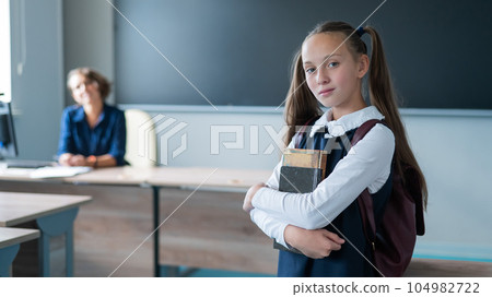 Caucasian girl and female teacher in the classroom. The schoolgirl is holding a textbook. 104982722