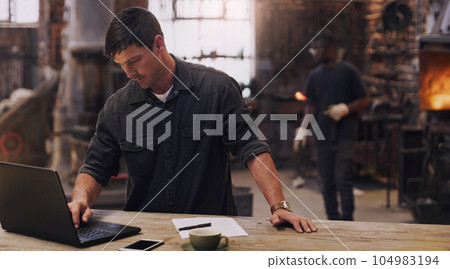 Workshop, man and working on laptop in metal blacksmith factory for trade business, finance and industry technology. Welder, computer and online paperwork for businessman, staff and forge worker 104983194