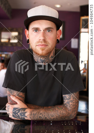 Tattoos, creative and portrait of a man with tattoos standing by the counter of his artistic studio. Confident, cool and punk man with a edgy, funky and body art or ink creativity business store. Tattoos, creative and portrait of a man with tattoos standing by the counter of his artistic studio. Confident, cool and punk man with a edgy, funky and body art or ink creativity business store. 104984735