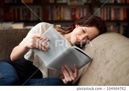 Relax, library and woman reading a book for literature while sitting on a sofa with shelves. Calm, happy and female person with a smile enjoying a fantasy story or novel on couch in bookstore or shop 104984762