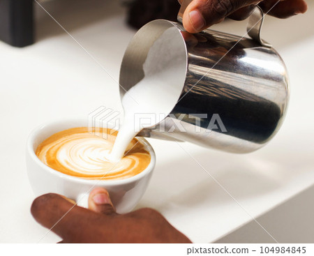 Coffee, closeup hand of a barista and pouring milk in a cup in a cafe in the morning. Latte or cappuccino, cafeteria and hands of a person pour a hot beverage for expresso drink at kitchen counter Coffee, closeup hand of a barista and pouring milk in a cup in a cafe in the morning. Latte or cappuccino, cafeteria and hands of a person pour a hot beverage for expresso drink at kitchen counter 104984845