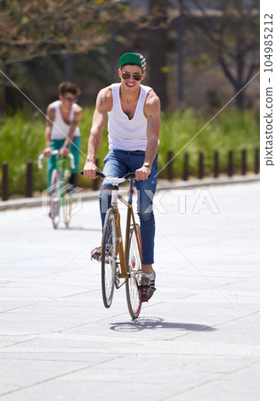 Men, summer and students in street, bicycle and sustainable transport for sunshine, freedom and wheelie. Young gen z people, retro bike and outdoor together in road for travel, university or campus Men, summer and students in street, bicycle and sustainable transport for sunshine, freedom and wheelie. Young gen z people, retro bike and outdoor together in road for travel, university or campus 104985212