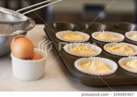 Cupcake tray, batter and baking in a kitchen with muffin mix or wrapper and cooking in a bakery. Closeup, paper cup and food with chef in a restaurant, coffee shop or cafe with sweet cake and pastry Cupcake tray, batter and baking in a kitchen with muffin mix or wrapper and cooking in a bakery. Closeup, paper cup and food with chef in a restaurant, coffee shop or cafe with sweet cake and pastry 104985407