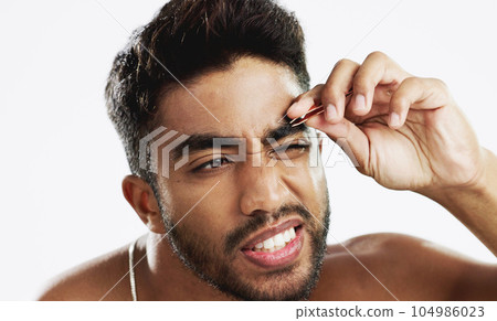 Man grooming his eyebrows with a tweezer in studio for self care, beauty and cleanliness. Hair removal, tweezing and male model from India doing facial epilation plucking routine by white background. Man grooming his eyebrows with a tweezer in studio for self care, beauty and cleanliness. Hair removal, tweezing and male model from India doing facial epilation plucking routine by white background. 104986023