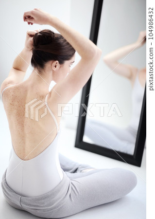 Female ballerina doing her hair by a mirror in a studio before a dance class, performance or concert. Art, creative and back of young ballet dancer getting ready for classical dancing in her room. 104986432